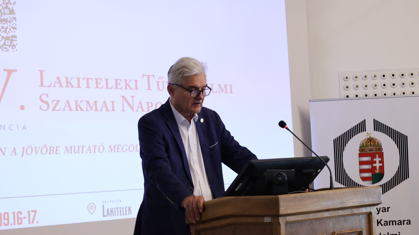 Előadó a konferencia pulpitusánál, a háttérben a rendezvény nyitódiája.